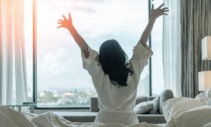 una donna a braccia alte in segno di gioia di schiena seduta su un letto guarda fuori da una grande finestra la città