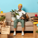 un uomo su un divano con una pianta e un cane in braccio e intorno degli scatoloni